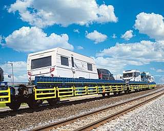 Ein Wohnwagen und ein Wohnmobil werden huckepack auf dem blauen AUTOZUG über den Sylt-Damm transportiert