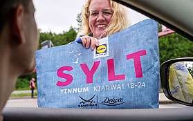 Eine Mitarbeiterin des blauen AUTOZUG Sylt reicht einem Kunden die kostenlose Sylttüüt ins Auto.
