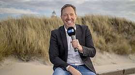 Presenter Frank Bremser sits with a microphone in front of a dune landscape on a wooden beam on the beach.