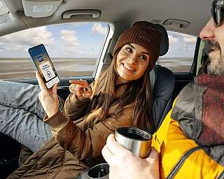 Ein Paar sitzt im Auto während der Überfahrt nach Sylt. Die Frau zeigt auf dem Handy den Stream des AUTOZUG RADIO Sylt.