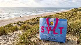 AI visual: The Sylttüüt of the blue AUTOZUG Sylt stands in the dunes in front of a dreamlike beach backdrop.